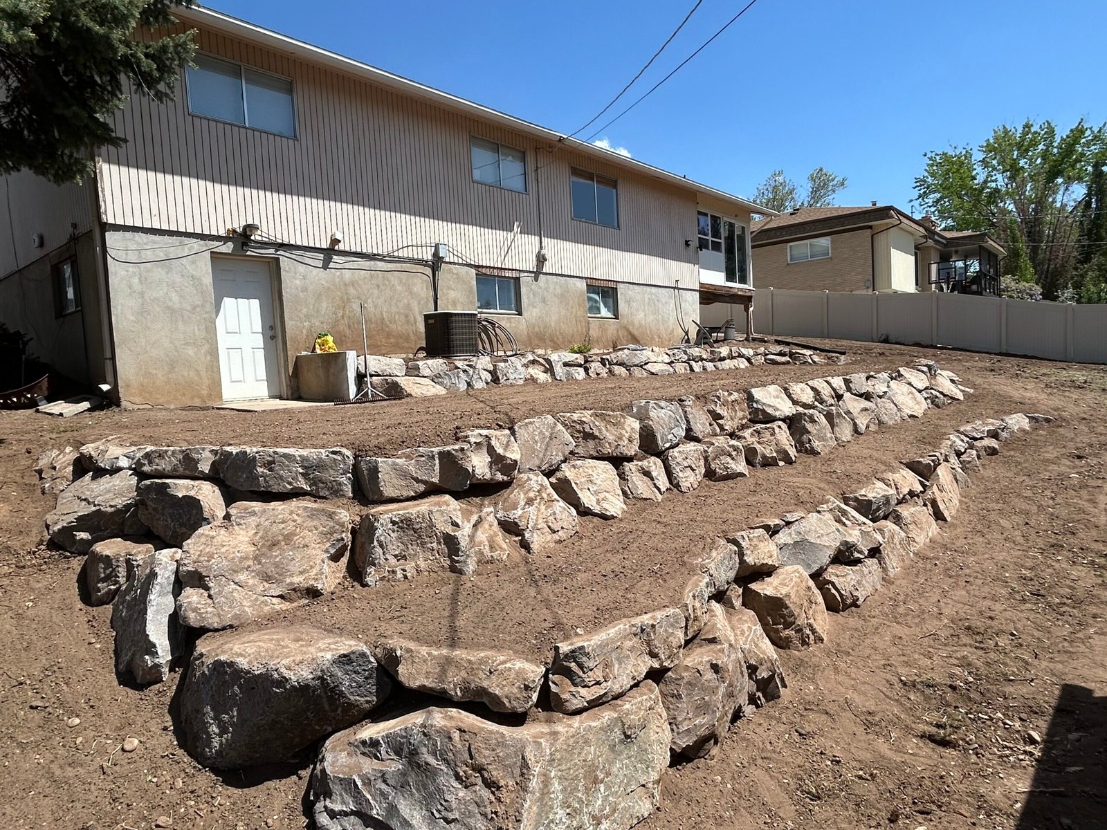 An image of a retaining wall behind a house. There are three tiers of elevation with two rows of rocks for each tier and fades into the hillside in the distance.